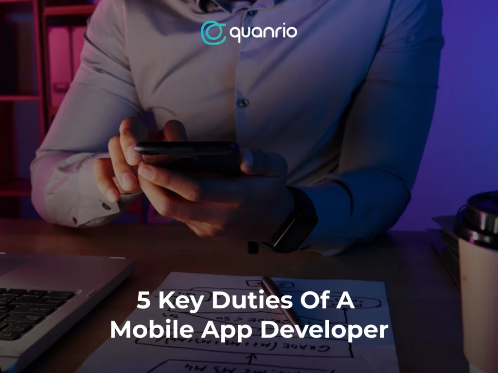 Person using a smartphone at a desk with a laptop, notepad, and coffee cup, with the text '5 Key Duties Of A Mobile App Developer' and the logo 'quanrio.